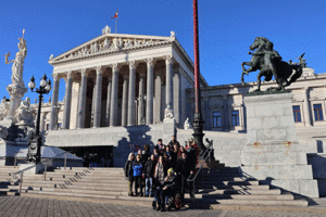 Gruppenbild. Sie befinden sich auf den Stufen vor dem Parlamentsgebäude. Im Hintergrund sieht man den überdachten Eingang mit mehreren Säulen. Rechts und Links hinter der Gruppe befindet sich je eine große Statue.