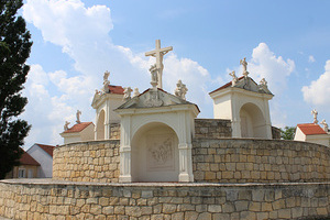 Ein steiniger Weg führt zu einer Kirchenfassade mit einem Kreuz und Statuen an der Spitze, umgeben von bewölktem Himmel.