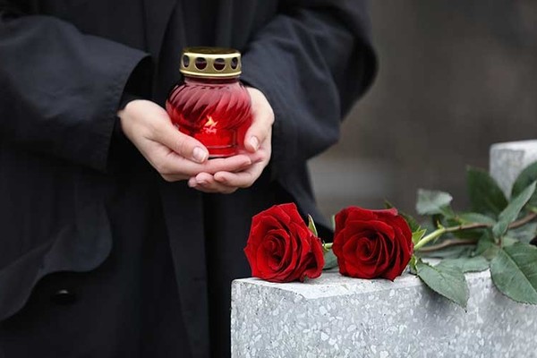 Woman holding candle near light grey tombstone with red roses outdoors, closeup. Funeral ceremony