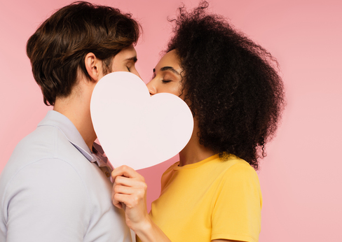 Dunkelhaariges Paar küsst sich, hält weißes Herz aus Karton vor die Münder