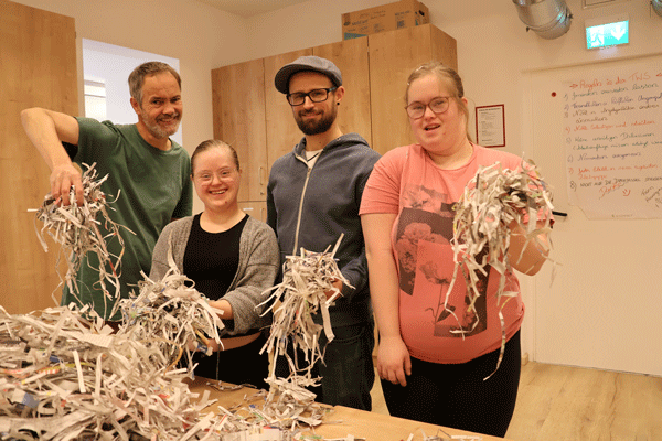Drei Klient*innen und ein Mitarbeiter der TWS Andreas mit Zeitungspapierschnipseln in den Händen und noch mehr Papier auf dem Tisch vor ihnen