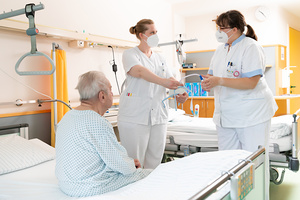 Das Foto zeigt einen Patienten, der von zwei Pflegemitarbeiterinnen versorgt wird.