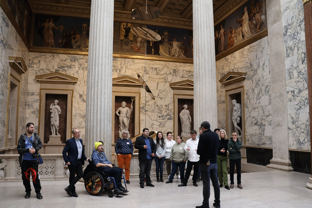 Die Gruppe befindet sich in einem Raum mit hohen Wänden aus Marmor. Zwei große Säulen sind im Hintergrund. An der Rückwand sind fünf Statuen in der Wand versenkt und ein Tourguide erklärt gerade etwas.