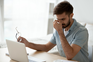 Mann in blauen Hemd sitzt vor Computer, mit einer Hand hält er seine Brille und mit der anderen Hand reibt er seine Augen.