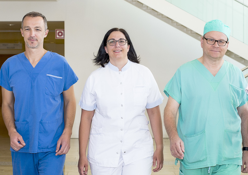 Das Foto zeigt einen Pfleger in dunkelblauer Arbeitskleidung, eine Pflegerin in weißer Arbeitskleidung und einen Pfleger in türkiser Arbeitskleidung