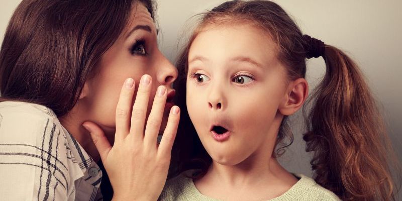 Shocked mother whispering the secret information to her surprising cute girl in ear on blue background
