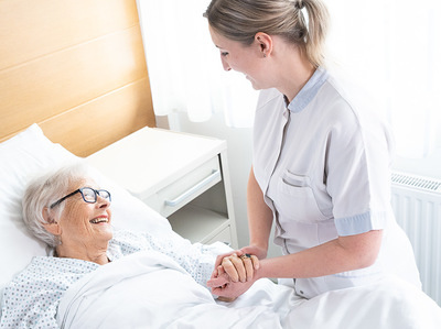 Das Bild zeigt eine ältere Patientin im Bett. Eine Pflegerin hält ihre Hand und sie lächeln sich an.