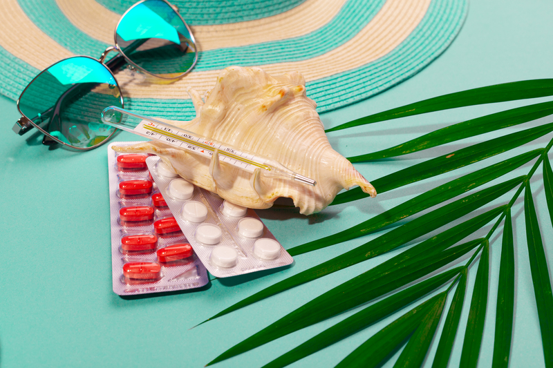 Top view on white and red fever pills and a thermometer in seashell, striped hat, sunglasses, blue flip-flops, palm leaf, on mint green background. Concept of preparing for sickness on vacation at sea. Advertising space.