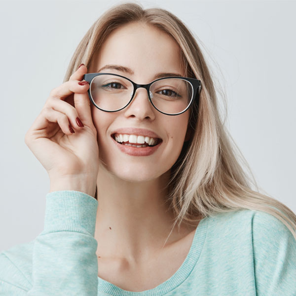 Das Foto zeigt eine Frau mit einer Brille, die in die Kamera lächelt und sich mit einer Hand auf die Brille greift.